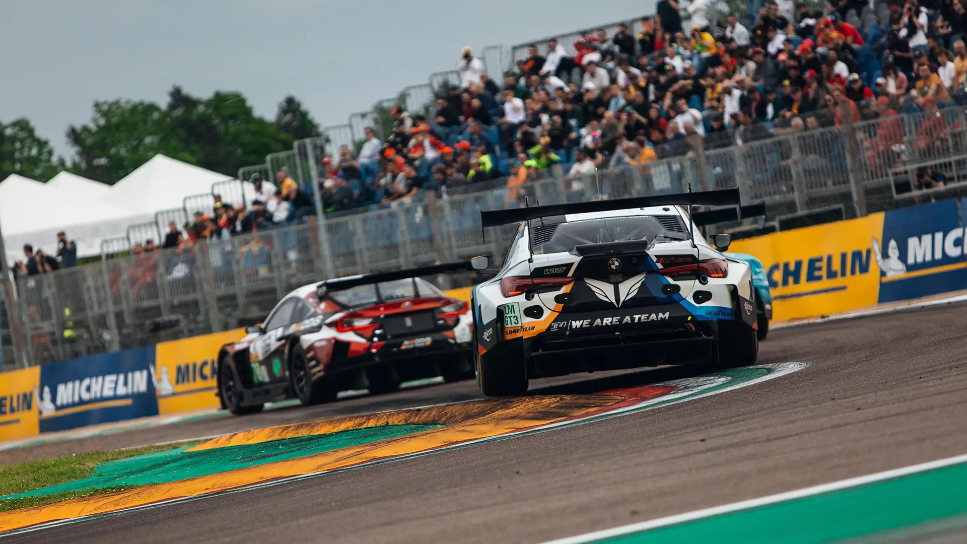 Two GT‑style race cars viewed from the rear as they corner on a racetrack, with spectators watching from grandstands in the background.