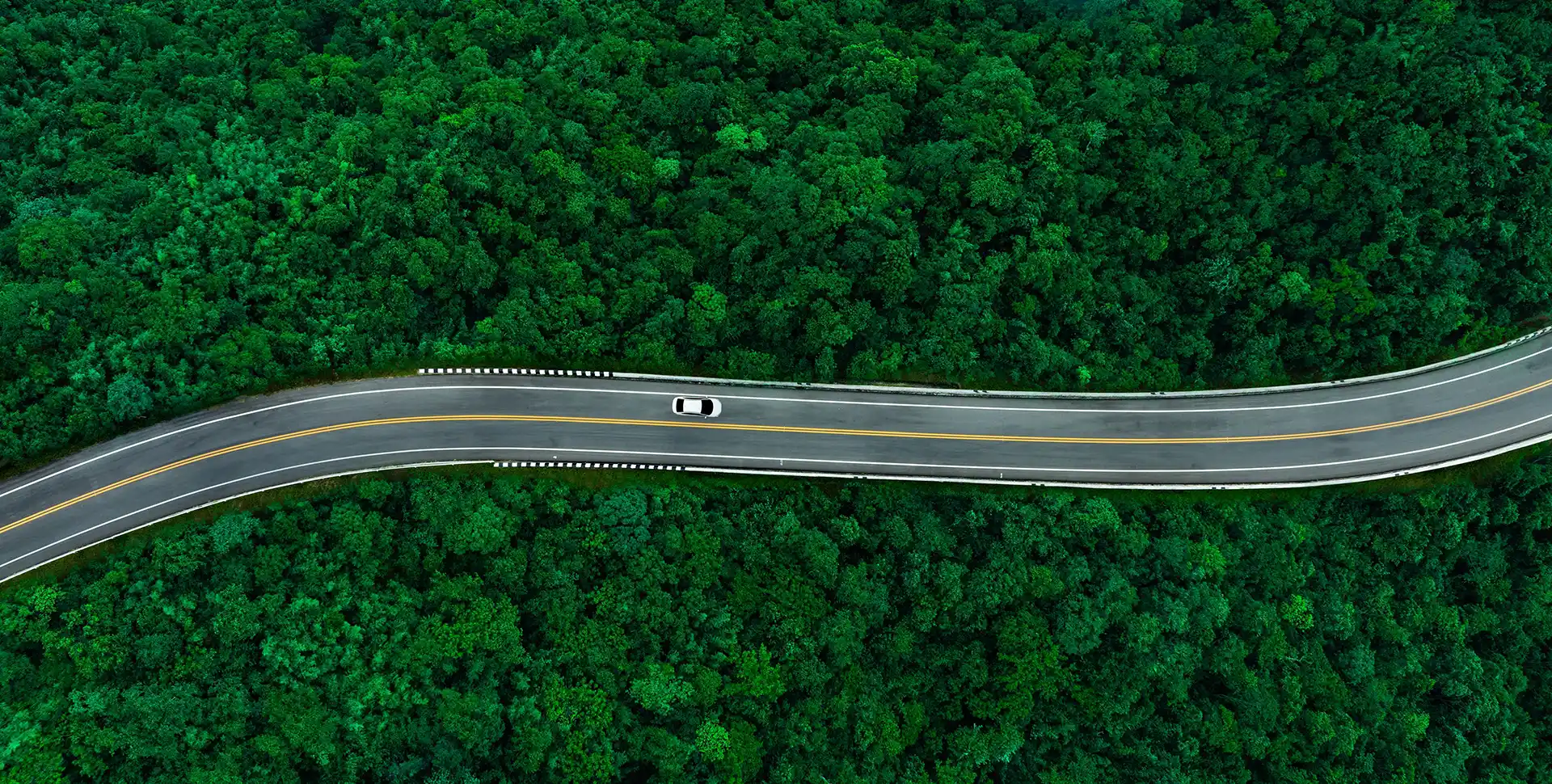 Aerial view of a winding two‑lane road cutting through dense green forest, with a single car driving along the curve.