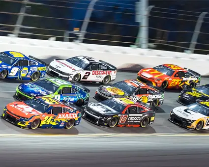 Pack of stock cars racing closely together on an oval track, shown side by side at speed with colorful liveries under stadium lighting.