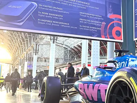 Formula‑style race car displayed indoors at an exhibition, viewed from the rear side, with people walking nearby and large overhead banners showing connectivity and performance information.