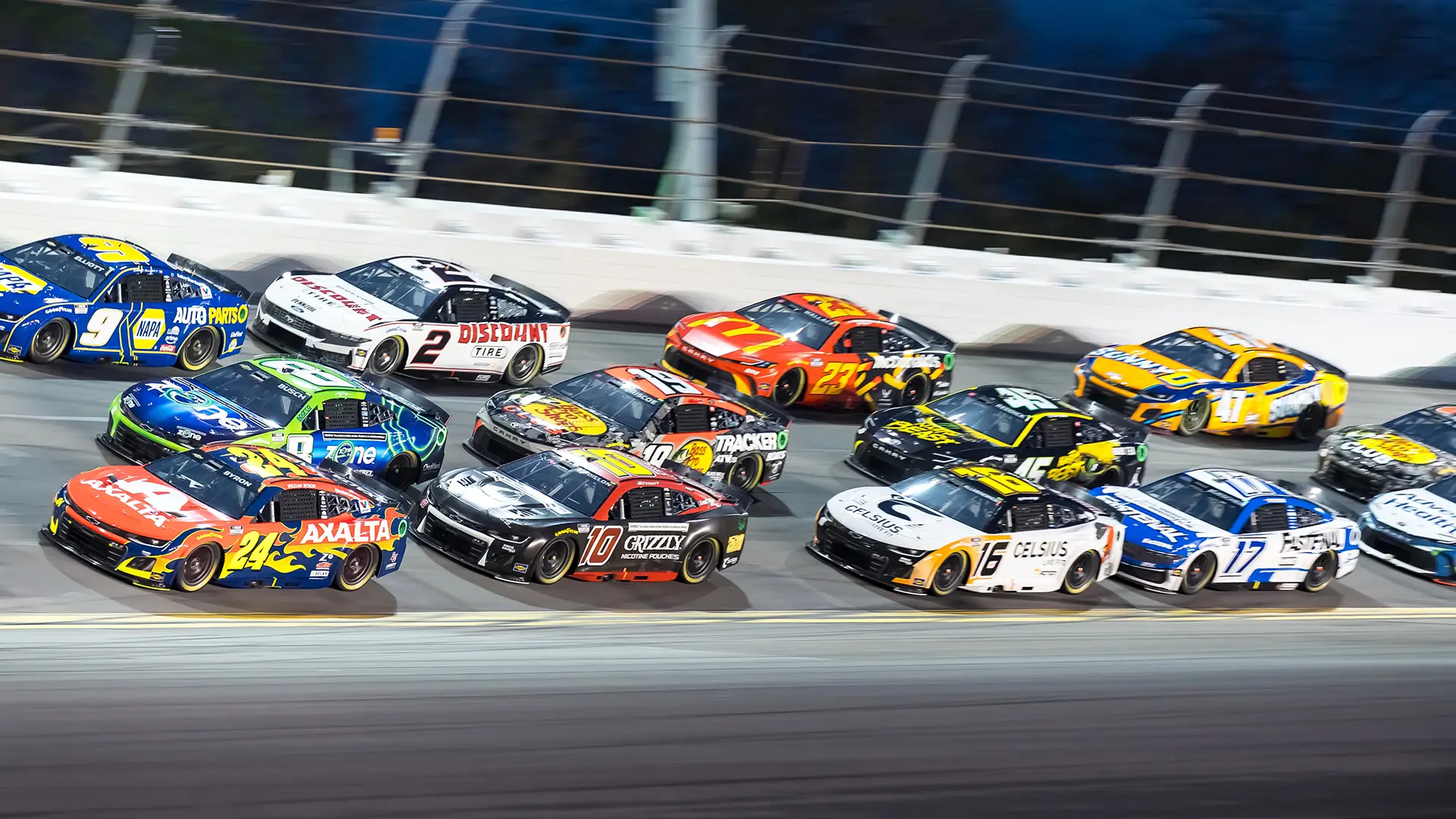 Pack of stock cars racing closely together on an oval track, shown side by side at speed with colorful liveries under stadium lighting.