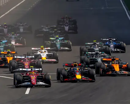 Group of Formula‑style race cars accelerating closely together on a racetrack, viewed from the front as they approach a straight section.